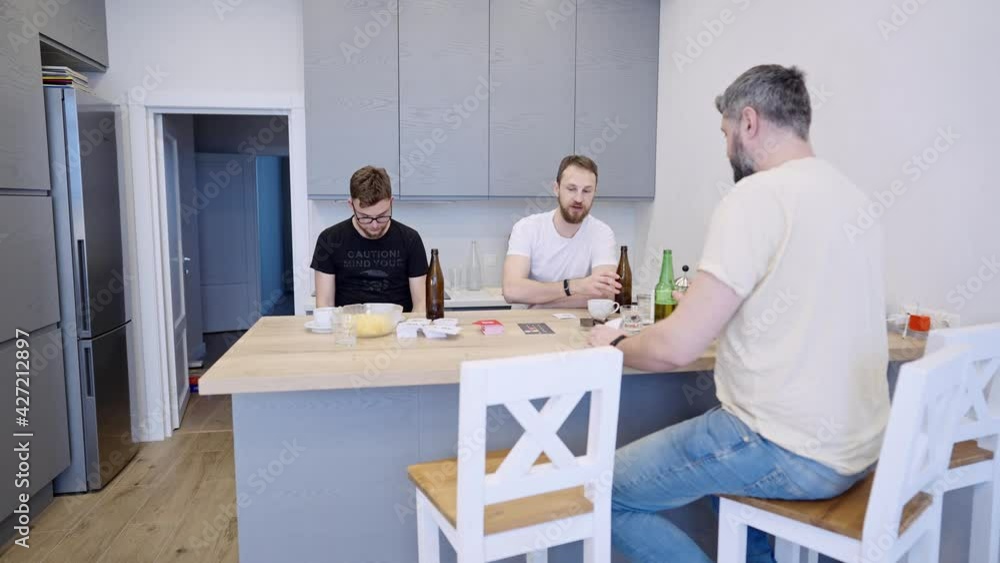 Three friends play board card games at home, sitting at the kitchen table. Fun communication and pastime with beer Three friends play board card games at home, sitting at the kitchen table. Fun communication and pastime with beer