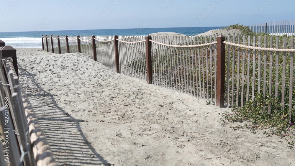 Pacific ocean coast, greenery and wooden picket fence on sea shore. Blue water waves on sunny summer beach, Encinitas shoreline, California USA. Coastline near Los Angeles. Coastal access entrance.