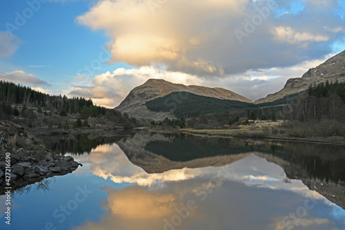 Sunset landscape of Argyll and Bute, Scotland with clouds reflected in small lochan, mountains and forest in background.