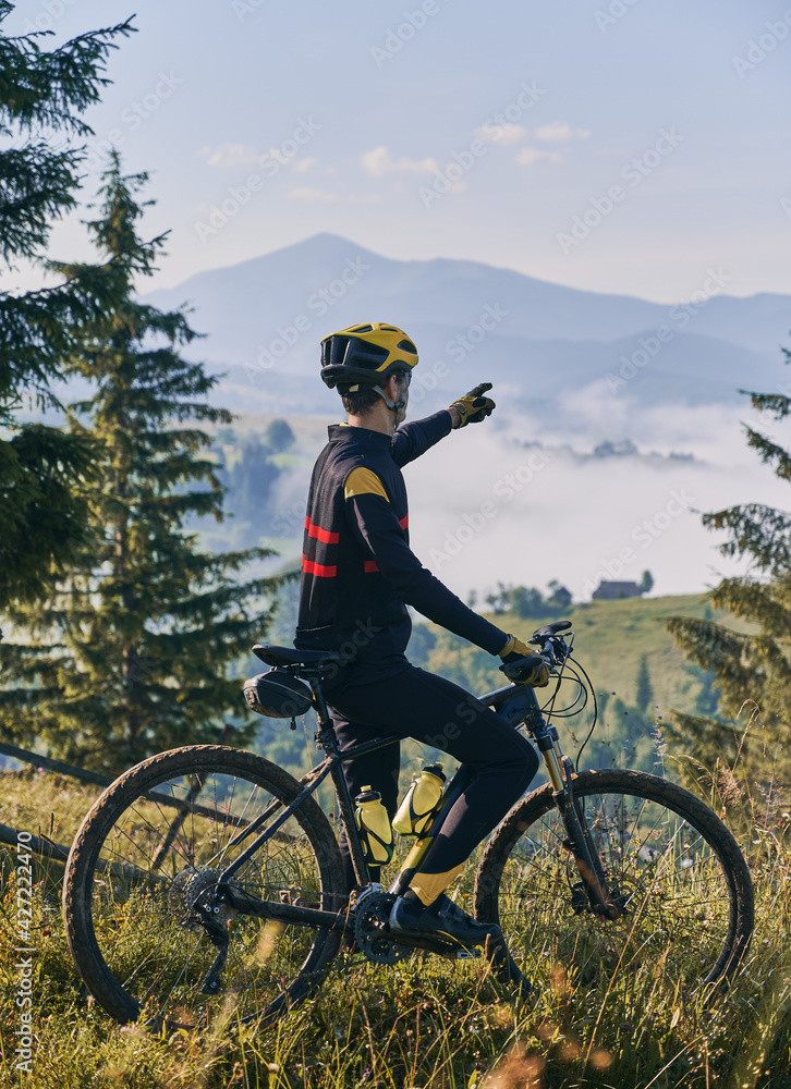 Obraz premium Back view of young man in cycling suit standing on grassy hill near bicycle and pointing finger at mountains. Male bicyclist in safety helmet standing with mountain bike. Concept of active leisure.