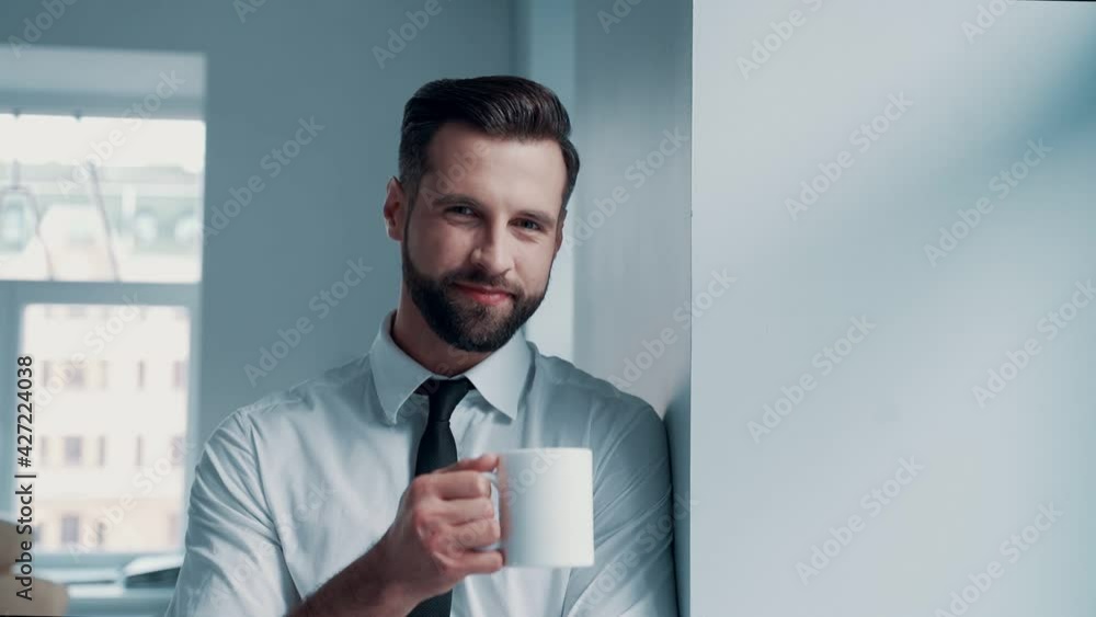 Cheerful young man in shirt and tie drinking hot drink and looking at camera while standing indoors Cheerful young man in shirt and tie drinking hot drink and looking at camera while standing indoors