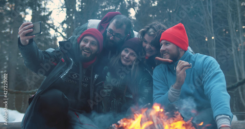 Young friends having outdoors barbeque in winter forest and taking selfie on smartphone