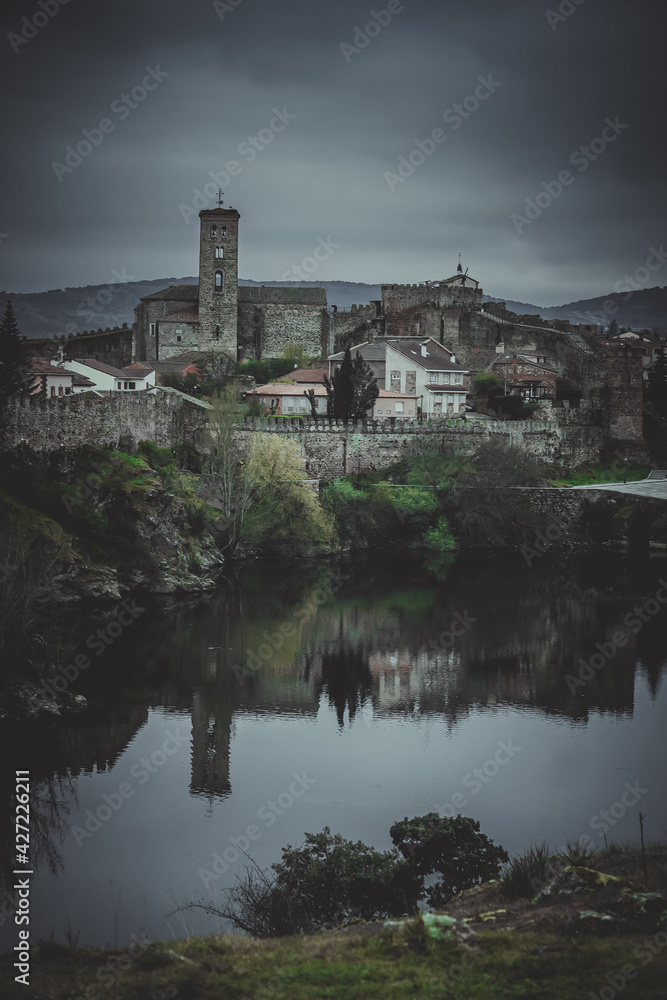 Romanesque village and church with city walls reflected in a lake with a cloudy sky and green areas with some trees.