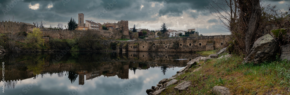 Panoramic view of a bridge and a village over a river with a cloudy sky and the village reflected in the water.