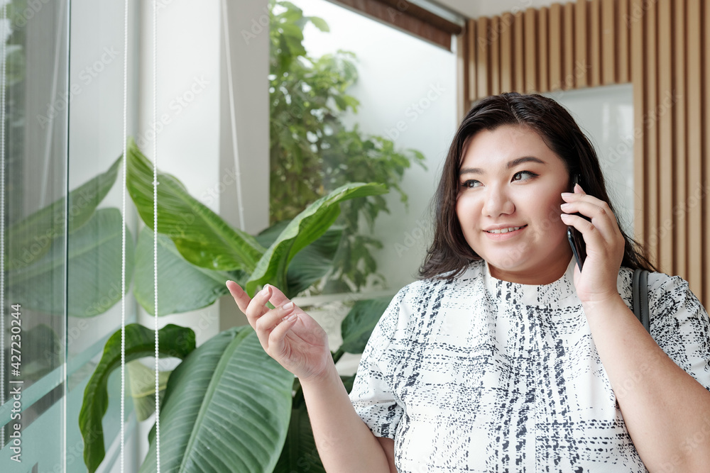 Pretty smiling young woman talking on phone with friend of colleague and looking through window of hotel or office building Pretty smiling young woman talking on phone with friend of colleague and looking through window of hotel or office building