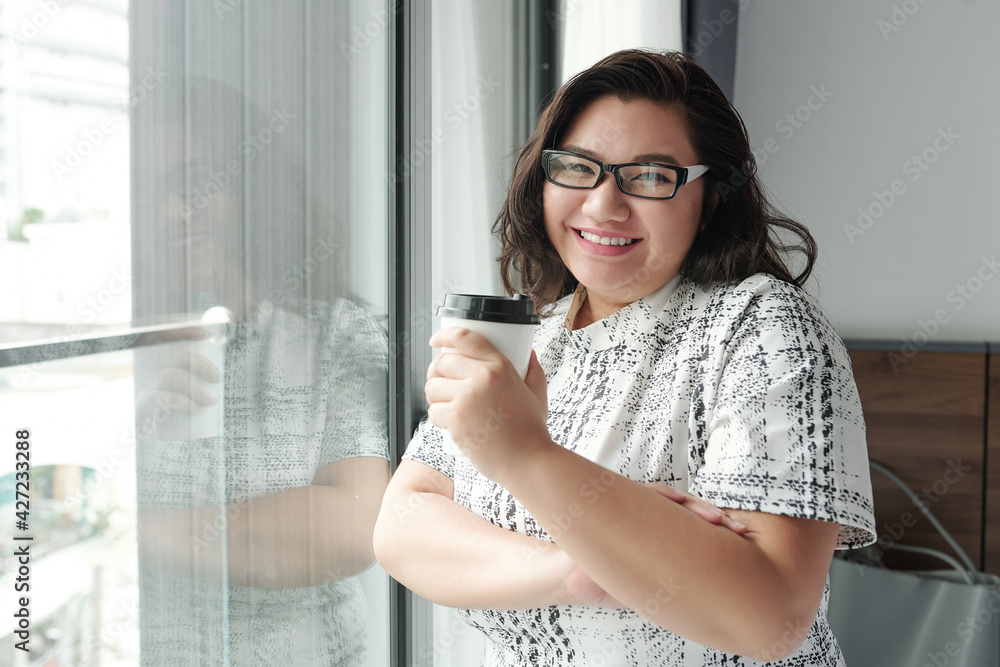 Portrait of happy young woman in glasses standing at hotel room window with cup of take out coffee in hands Portrait of happy young woman in glasses standing at hotel room window with cup of take out coffee in hands