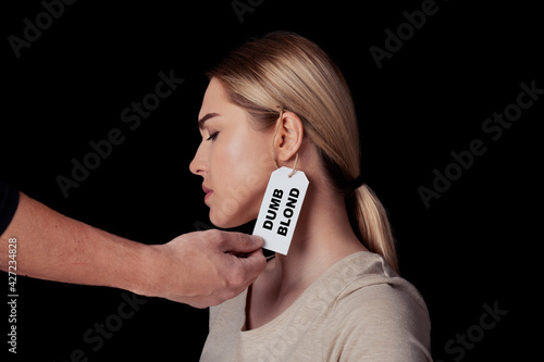 Hand of man touch face of upset young woman with label on ear is dumb blonde