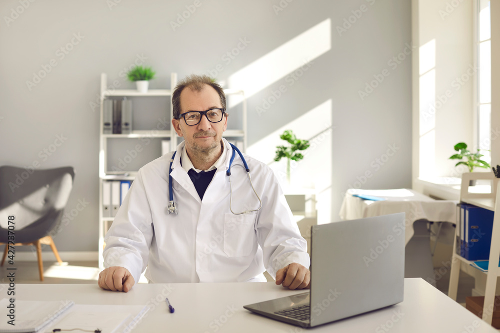 Doctor man wearing white coat with stethoscope sitting at desk with laptop in hospital office looking at camera headshot portrait. Perfect medical service, telemedicine, remote healthcare, insurance Doctor man wearing white coat with stethoscope sitting at desk with laptop in hospital office looking at camera headshot portrait. Perfect medical service, telemedicine, remote healthcare, insurance