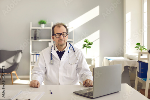 Doctor man wearing white coat with stethoscope sitting at desk with laptop in hospital office looking at camera headshot portrait. Perfect medical service, telemedicine, remote healthcare, insurance