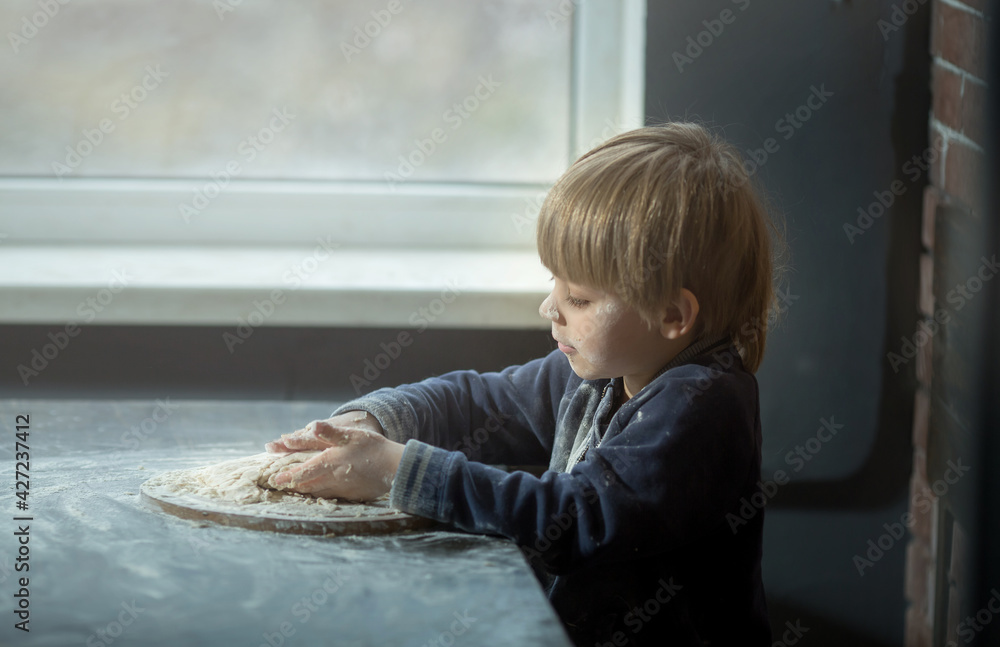 Fototapeta premium a small child boy kneads the dough, he is all soiled in flour, helps his mother to cook. Brick and wood background