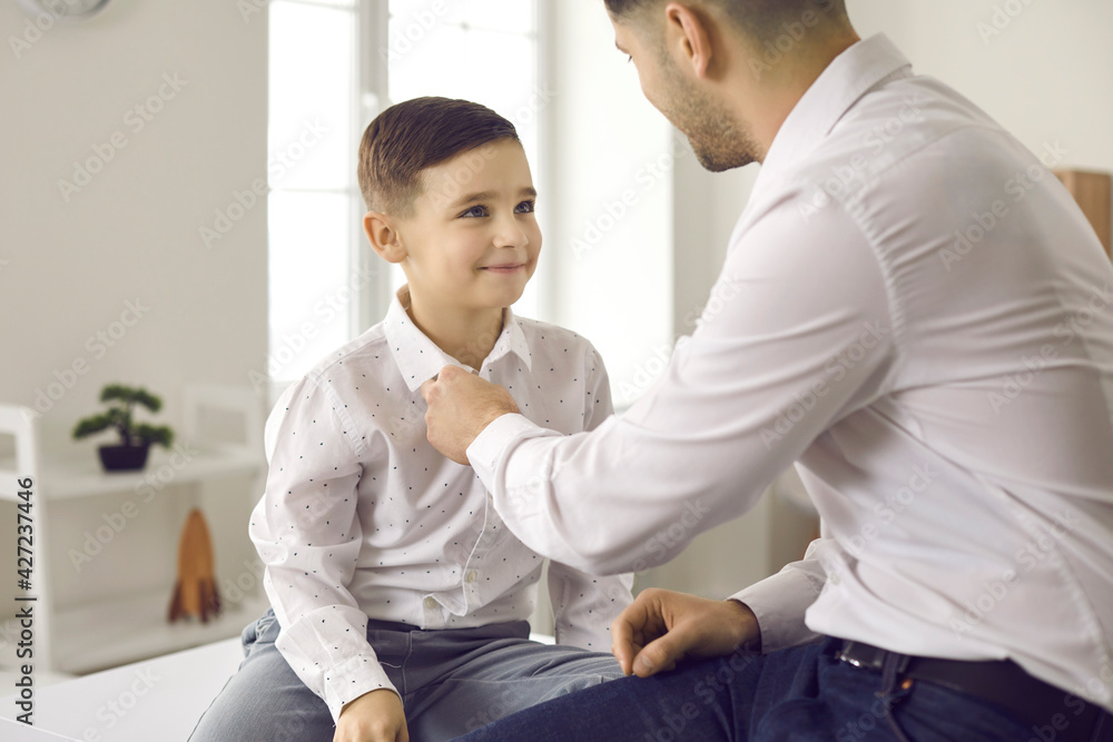 Dad as a best friend and mentor. Loving father helps his cute little son to fasten a button on his shirt. Boy wants to look like his handsome father who wears jeans and a shirt. Stylish family. Dad as a best friend and mentor. Loving father helps his cute little son to fasten a button on his shirt. Boy wants to look like his handsome father who wears jeans and a shirt. Stylish family.