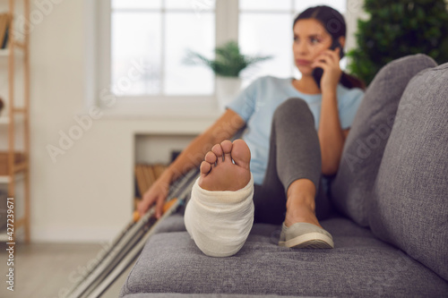 Fotografia Young woman with broken leg in cast lying relaxing on sofa with crutches nearby and talking on phone at home, selective focus