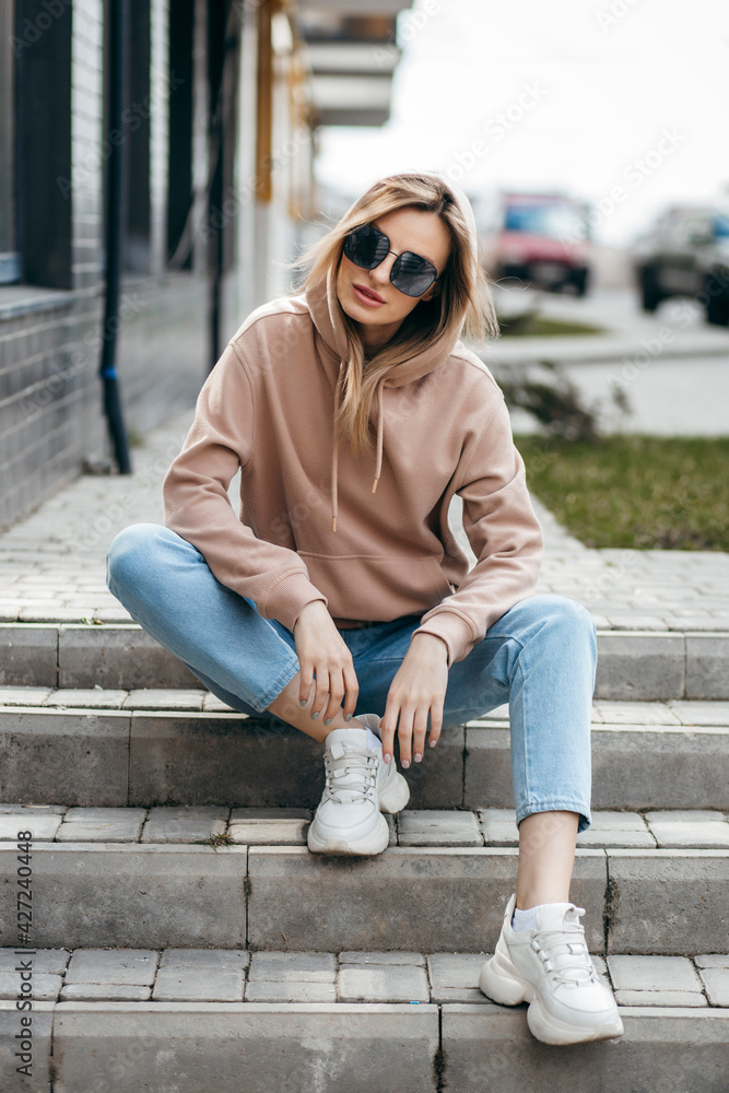 Fasion blonde woman in brown oversize hoodie, glasses and blue jeans posing sitting on the steps.  Mockup for logo or branding design