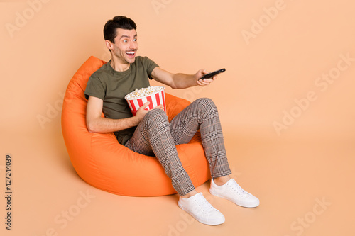 Full size profile photo of impressed brunet guy sit watch tv wear t-shirt trousers sneakers isolated on beige color background