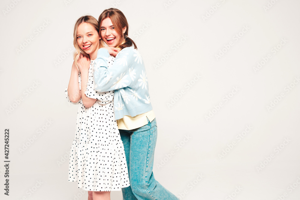 Two young beautiful blond smiling hipster female in trendy summer clothes. Sexy carefree women posing on grey background in studio. Trendy and positive models having fun Two young beautiful blond smiling hipster female in trendy summer clothes. Sexy carefree women posing on grey background in studio. Trendy and positive models having fun