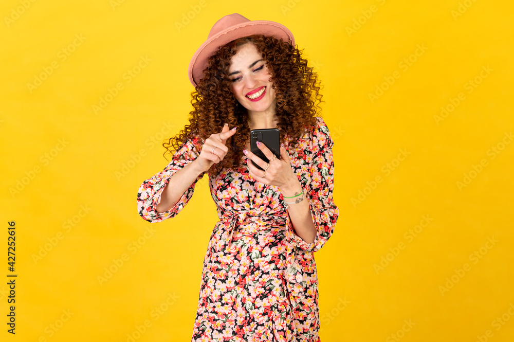 Girl in summer outfit with curly hair using smartphone and making facial expressions. Isolated on yellow background. Scrolling on the phone screen. Girl in summer outfit with curly hair using smartphone and making facial expressions. Isolated on yellow background. Scrolling on the phone screen.