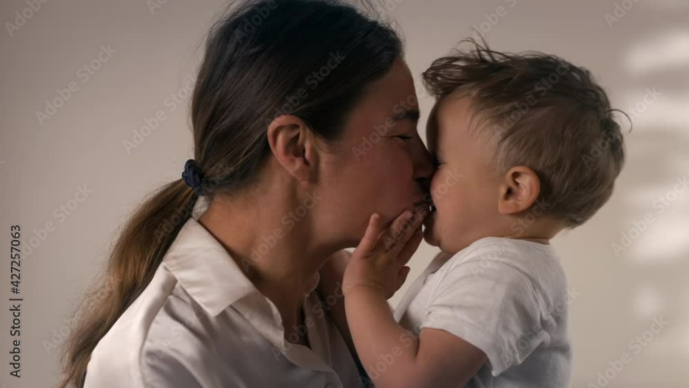 Authentic shot of young happy mother is kissing and playing with her toddler baby boy while keeping he in her arms in a nursery. Concept of love, parenthood, childhood, life, maternity, motherhood. Authentic shot of young happy mother is kissing and playing with her toddler baby boy while keeping he in her arms in a nursery. Concept of love, parenthood, childhood, life, maternity, motherhood.