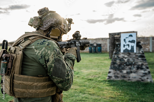 Soldier in the uniform (Cropat woodland pattern) taking aim at the target on the shooting range with his personal weapon. He is wearing ballistic helmet and plate carrier in coyote brown.