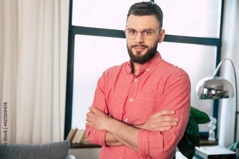Handsome confident and happy bearded man in eyeglasses and shirt is standing with crossed arms and looking on the camera Handsome confident and happy bearded man in eyeglasses and shirt is standing with crossed arms and looking on the camera