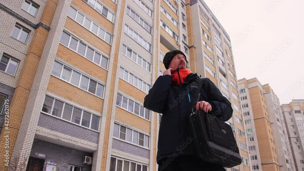 A business man with a beard and mustache stands with a bag on his shoulder. Male in a warm jacket on a city street in cool weather. In the background, multi-storey residential buildings out of focus. A business man with a beard and mustache stands with a bag on his shoulder. Male in a warm jacket on a city street in cool weather. In the background, multi-storey residential buildings out of focus.