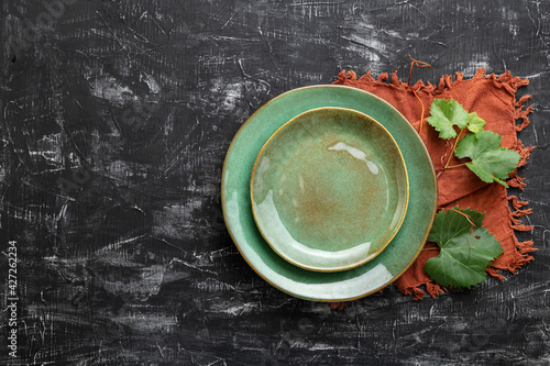 Empty green plate served with grape leaf plant and table napkin. Mockup template plate for luxury dinner in wine restaurant with Mediterranean cuisine. Copy space on dark black concrete table top view