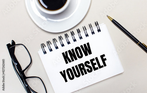 Glasses, a white cup of coffee, a white notebook with the words KNOW YOURSELF and a ballpoint pen lie on a light background. Flat lay. View from above.