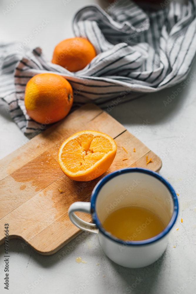Half cut orange on wooden board, freshly squeezed into retro enamel mug, organic oranges and kitchen cloth on white table. Making juice from citrus fruit. Healthy diet lifestyle. Selective focus. Half cut orange on wooden board, freshly squeezed into retro enamel mug, organic oranges and kitchen cloth on white table. Making juice from citrus fruit. Healthy diet lifestyle. Selective focus.