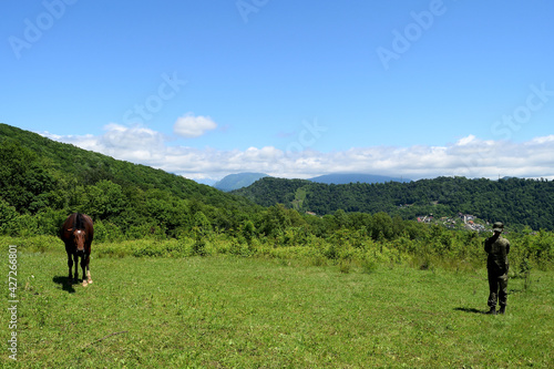 high blue sky with rare clouds. mountains with green trees that a man looks at, a field with low dense grass that a horse eats.
