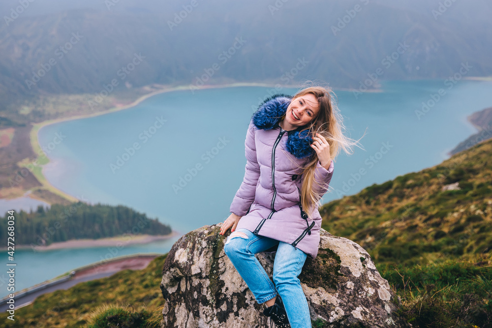 Naklejka premium a girl sits on a mountain, the wind develops her hair, A landscape view of the Lagoa do Fogo caldera under a foggy sky with great depth.
