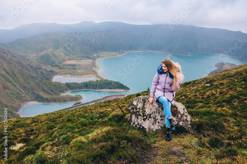 A woman sits on a stone against the backdrop of the beautiful crater of Lake Lagoa do Fogo in the Agua de Pau Massiva stratovolcano in the center of São Miguel Island. Travel to the Azores.