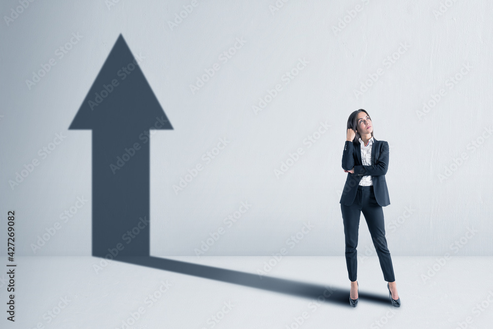 Fototapeta premium Businesswoman with a shadow in the shape of an arrow up on the blank white concrete wall background. Promotion and success concept