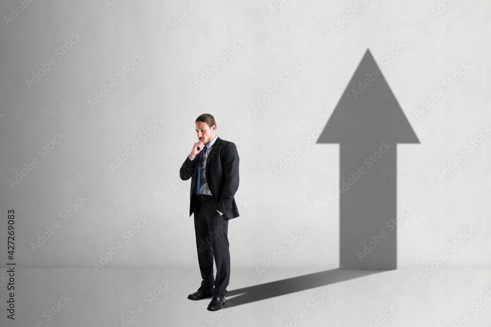 Businessman with a shadow in the shape of an arrow up on the blank white concrete wall background. promotion and success concept Businessman with a shadow in the shape of an arrow up on the blank white concrete wall background. promotion and success concept