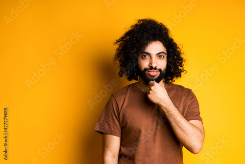Smiling african american guy with a beard and in a brown t-shirt touches his beard while standing on a yellow background