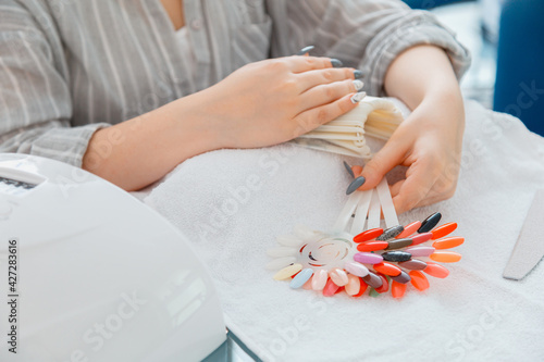 Female hands with artificial acrylic nails picks up new nail polish color during manicure procedure. Colored nail polish process on manicure in beauty salon. Hygiene and beauty of hands in nail salon