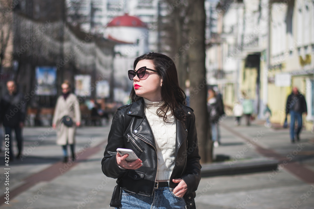 Fototapeta premium Young cheerful stylish woman using cell phone and texting message on city street