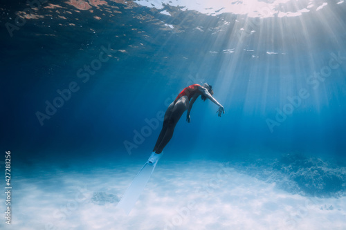 Woman freediver in swimsuit with fins glides underwater over sand in ocean.