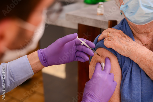 elderly senior woman with surgical mask having covid 19 vaccine injection by doctor male nurse at home