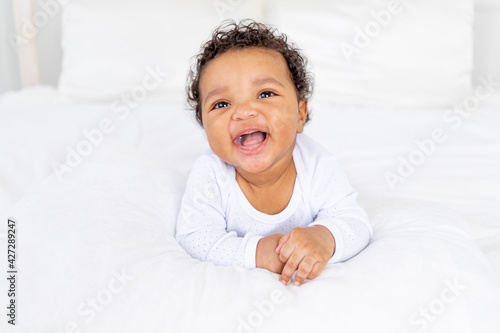 smiling African-American little baby on white bed in bedroom laughing