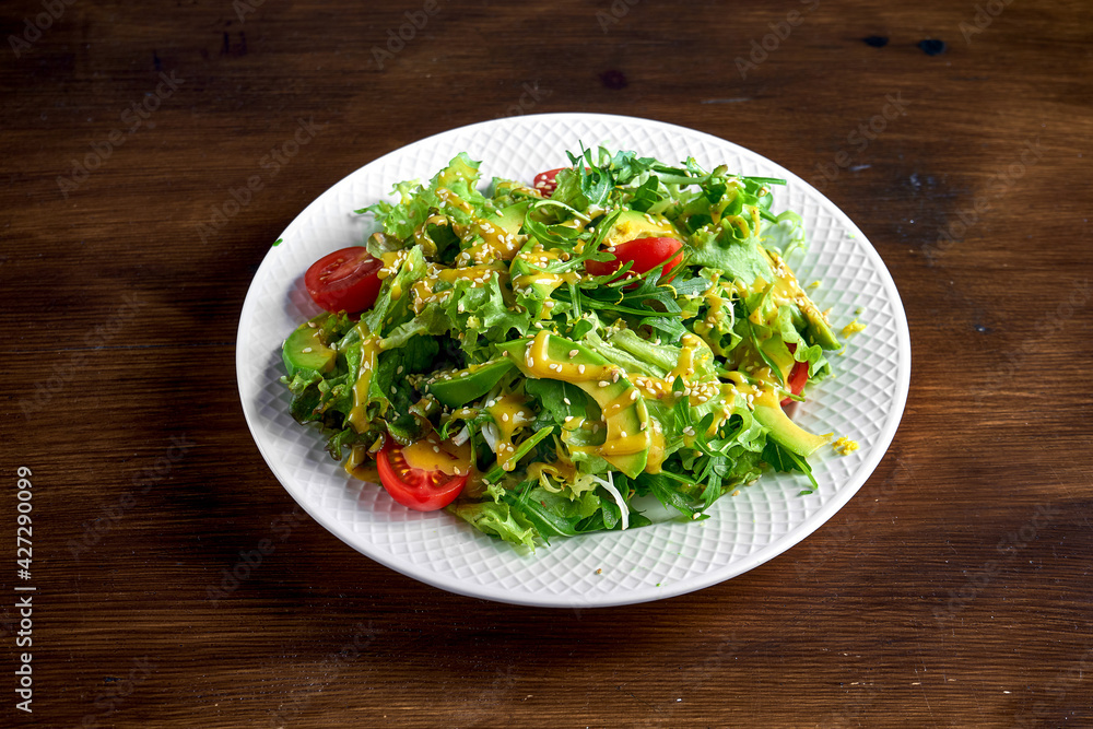 Healthy and dietary salad mix with avocado, sesame seeds, yellow dressing and cherry tomatoes, served in a white plate on a wooden background
