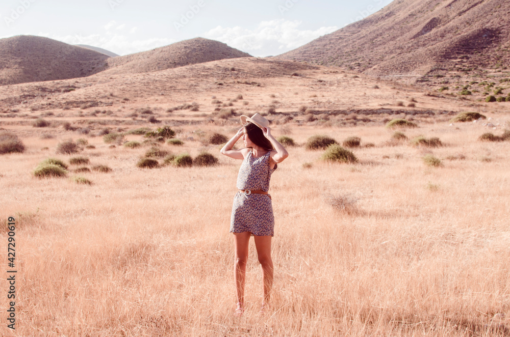 Young woman with long brunette hair holding a hat wears a floral dress in a dry meadow in the late afternoon