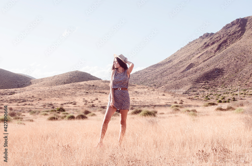 Fototapeta premium Young woman with long brunette hair holding a hat wears a floral dress in a dry meadow in the late afternoon