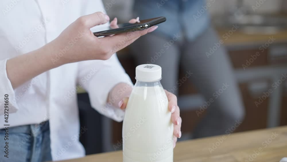 Close-up of unrecognizable woman scanning QR code on milk bottle with smartphone. Young slim lady using modern technologies in everyday life in kitchen at home Close-up of unrecognizable woman scanning QR code on milk bottle with smartphone. Young slim lady using modern technologies in everyday life in kitchen at home