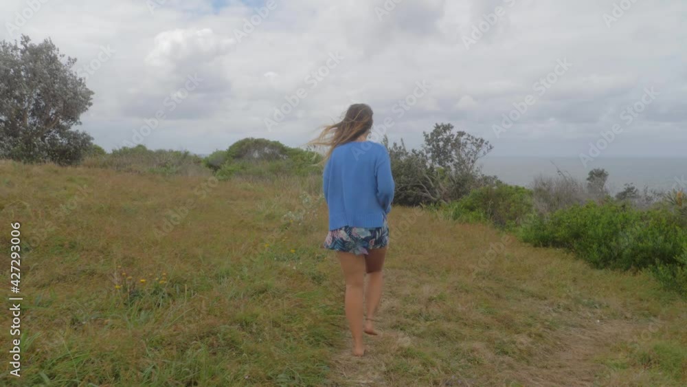 Beautiful Caucasian Woman Walking In Bare Feet On Grassy Hill By The Sea Coast. Big Nobby At Crescent Head Lookout In New South Wales, Australia. rear shot Beautiful Caucasian Woman Walking In Bare Feet On Grassy Hill By The Sea Coast. Big Nobby At Crescent Head Lookout In New South Wales, Australia. rear shot