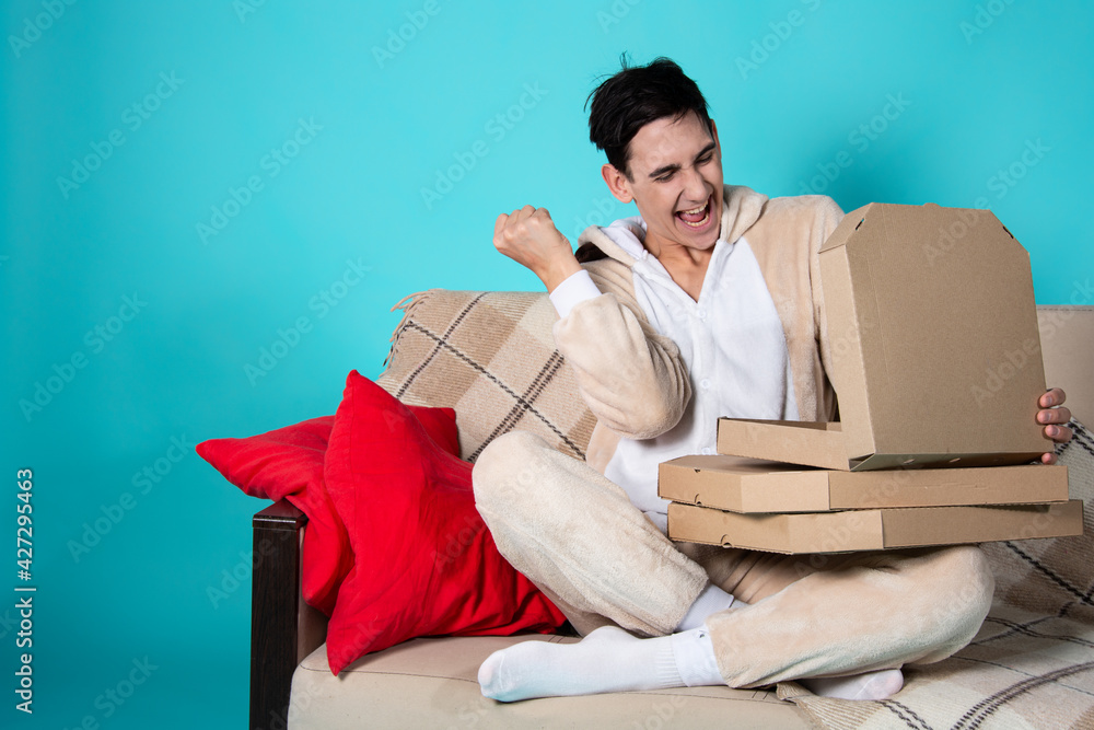 A cute young man in pajamas sits on the couch with a box of pizza. Beige sofa and red pillows. Rest in the evening with food. Student life. Blue background.