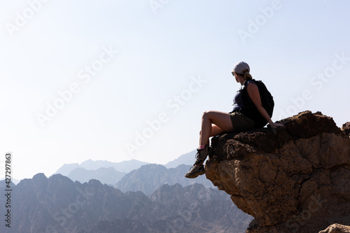 Young female tourist girl sitting at the edge of the rock with abyss below, looking at the mountain ridges in the horizon, Hatta, Hajar Mountains, United Arab Emirates.