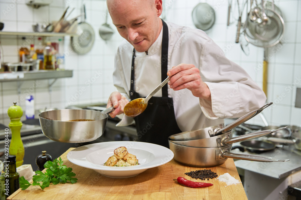 Chef without hair prepares scampi in the kitchen. He holds a pot in his hand and has a spoon in his hand. In the foreground you can see chily, pepper, salt, parsley. Feel like going out for dinner? Chef without hair prepares scampi in the kitchen. He holds a pot in his hand and has a spoon in his hand. In the foreground you can see chily, pepper, salt, parsley. Feel like going out for dinner?