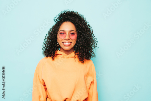 Beautiful black woman with afro curls hairstyle.Smiling model in orange hoodie and trendy jeans clothes. Sexy carefree female posing near blue wall in studio in sunglasses