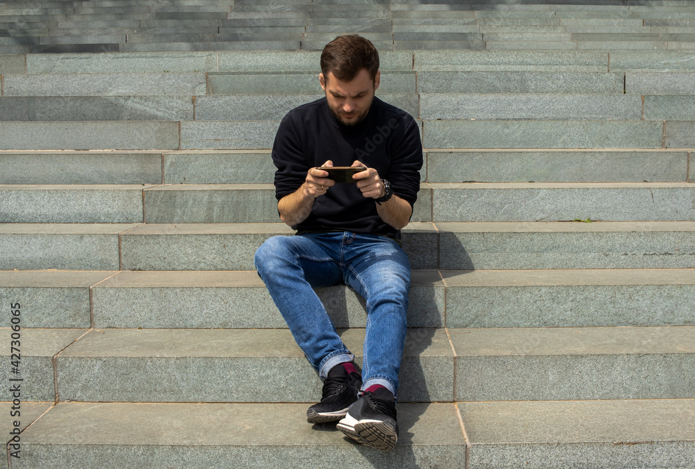 Young handsome man with a smartphone in his hands sitting on stone stairs and typing the message. Brutal man wearing jeans and sweatshirt using his mobile phone. People and social network concept. Young handsome man with a smartphone in his hands sitting on stone stairs and typing the message. Brutal man wearing jeans and sweatshirt using his mobile phone. People and social network concept.