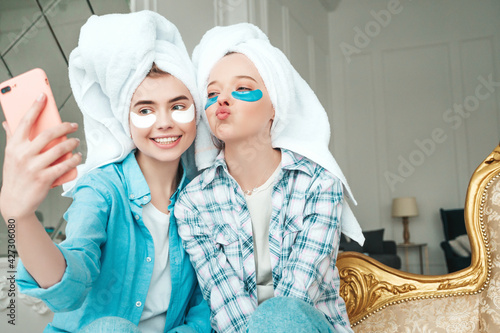 Two young beautiful smiling women with patches under their eyes. Sexy carefree models sitting on bed in posh apartment or hotel room. They doing beauty treatments at home in towels on heads