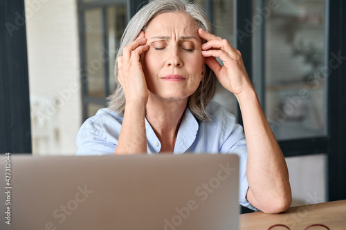Stressed middle 60s aged worker woman massaging head suffering of headache in home office. Senior old sick lady sitting at table on computer feeling overworked burned or menopause health disease.